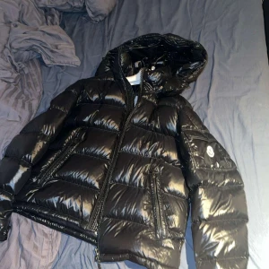 Moncler Maya - Säljer en svart, glansig pufferjacka från Moncler med huva och dragkedja. Jackan har Moncler-logga på ärmen, stora fickor med dragkedja och en cool serietidningsetikett på insidan. Perfekt för kalla dagar när du vill hålla stilen.            Äkta såklart, har kvitto från nk, kan mötas i Stockholm eller skickas med frakt
