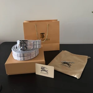 Burberry rutigt bälte med B-spänne - Snyggt bälte från Burberry med klassiskt rutigt mönster i beige, grått och vinrött. Bältet har ett stilrent metallspänne med Burberrys ikoniska B-logga. Perfekt accessoar för att lyfta din outfit med en lyxig touch.