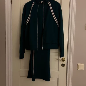 Mörkgrön set  med sportiga ränder - Mörkgrön set  i mjukt, stretchigt material med vita och mörkblå sportiga ränder längs sidorna. Byxorna har en utsvängd passform och är perfekt för en avslappnad och trendig look. Materialet känns som polyester och har en lätt glans. Den är nopprig på insidalår, vid buken och ungefär vid bröstet på zip tröjan