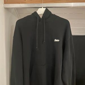 Svart pull&bear hoodie med tryck - Svart hoodie från SWTP med vit logga på bröstet och stort tryck på ryggen. Klassisk känguruficka, huva med snörning och mjukt material. Perfekt för dig som gillar streetwear och enkel stil.