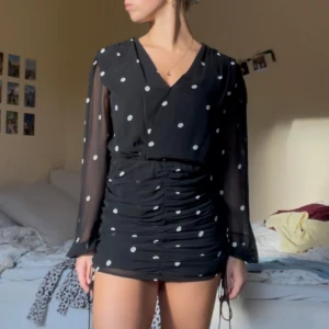 Svart prickig klänning - Svart kort klänning med vita prickar, långa transparenta ärmar och rynkade detaljer framtill. Klänningen har v-ringning och snörning i sidorna för en snygg passform.