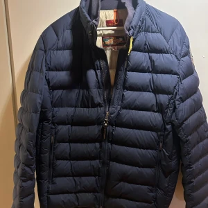Mörkblå dunjacka från Parajumpers XL - Säljer en mörkblå dunjacka från Parajumpers i storlek XL. Jackan har quiltad design, hög krage och dragkedja framtill samt på fickorna. Perfekt för kyliga dagar och har en klassisk, sportig look.
