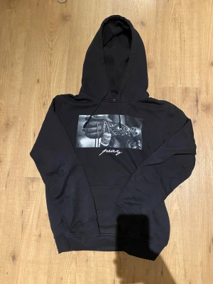 Svart hoodie Mister Tee pray L - Svart hoodie från Mister Tee i storlek L med tryck på bröstet som visar en hand med tatueringar och halskedjor, samt texten 'pray' under motivet. Tröjan har huva med snörning och är långärmad. Materialet är mjuk bomull, perfekt för chill dagar.