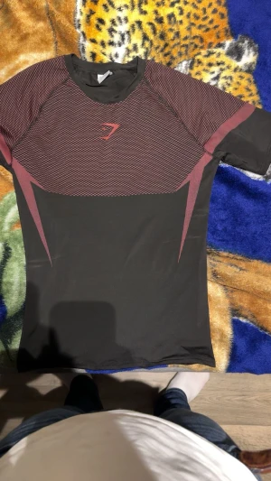 Svart och vinröd Gymshark t-shirt - Säljer en snygg svart och vinröd t-shirt från Gymshark med korta ärmar. T-shirten har ett grafiskt mönster i vinrött över bröst och rygg samt Gymshark-logga framtill. Materialet är stretchigt och andas, perfekt för träning. Passformen är tight och sportig.