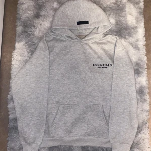 Grå Essentials Fear of God hoodie - Säljer en grå hoodie från Essentials Fear of God med svart logga på bröstet och ryggen. Tröjan har huva, känguruficka och är långärmad. Perfekt för dig som gillar streetwear och vill ha en clean look. Inte polyester utan materialet är Bomull och passar för de som är 180~185 cm lång 
