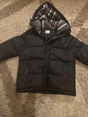 Svart pufferjacka från Burberry | Skicka prisförslag  - Snygg svart pufferjacka som har ärmar som går att ta bort så att de kan bli en väst från Burberry med huva och det ikoniska rutiga fodret i grått, vitt och rött. Jackan har två fickor med dragkedja, quiltad design och en Burberry London England-patch på ena ärmen som ger en exklusiv look. Jättevarm, bekväm och perfekt för kalla dagar. Passar både till streetstil och mer uppklätt. I fint skick och redo för en ny ägare.                              📏 Storlek: S ✨ Skick: Mycket bra 📦 Skickas snabbt