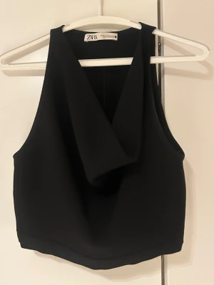 Svart croppad topp från Zara - Säljer en svart croppad topp från Zara i storlek XS. Toppen har en snygg omlott-detalj i halterneck vibe!