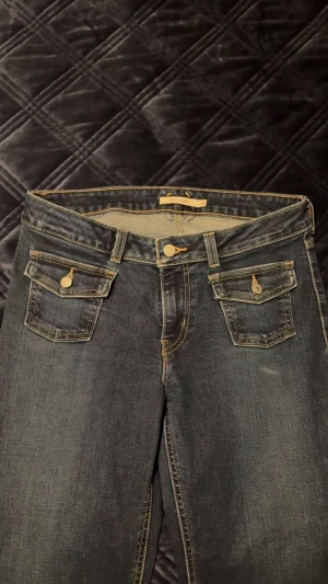 Levi's 501 mörkblå jeans, storlek 28 - Snygga Levi's 501 jeans i mörkblå tvätt med klassiska fem fickor och gul kontrastsöm. Modellen är bootcut och låg passform, med knappar och detaljer på fickorna. Perfekt för dig som gillar tidlös denimstil.