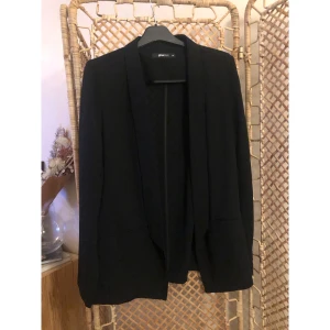 Svart kavaj från Gina Tricot - Stilren svart kavaj från Gina Tricot i storlek 44. Klassisk rak modell med smala slag och två fickor framtill. Perfekt att slänga över en t-shirt eller skjorta för en snygg look. Materialet känns mjukt och har en lätt struktur. Använd en gång. 