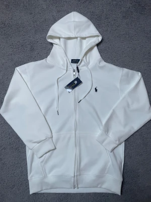 Vit hoodie från Polo Ralph Lauren  - Vit hoodie från Polo Ralph Lauren i storlek M men den sitter litte mindre. Tröjan har huva med snörning, hel dragkedja och klassisk broderad logga på bröstet. Tillverkad i mjuk bomull med ribbade muddar och fickor framtill. 