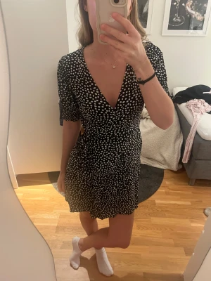 Svart prickig omlottklänning - Snygg svart kortklänning med vita prickar och omlottmodell. Klänningen har v-ringad hals, knyt i midjan och korta puffärmar. Den är lätt och flowy, perfekt för dig som gillar en feminin och trendig look.