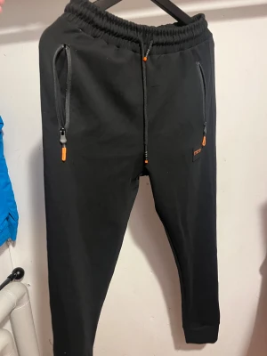 Svarta mjukisbyxor från BOSS - Svarta mjukisbyxor från BOSS med orange detaljer på snörning och dragkedjor. Byxorna har elastisk midja, smal passform och ribbade muddar vid benslut samt att materialet är mjukt och bekvämt, 