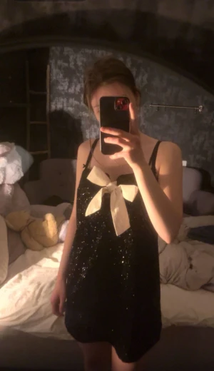 Svart glittrig kortklänning med rosett storlek S  - Supersöt svart glittrig kortklänning med smala axelband och stor vit rosett framtill. Klänningen har en rak passform och är perfekt för dig som vill sticka ut. Materialet är glittrigt och skimrar snyggt i ljuset. Ingen synlig logga eller märke. Storlek S, jag är 160. 