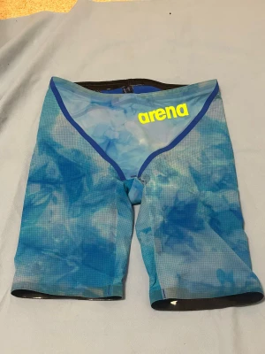 Blå Powerskin carbon glide från Arena - Säljer ett par blå Powerskin badshorts från Arena med coolt mönster och gul logga framtill. Shortsen har midwaist passform och är gjorda i syntetmaterial som torkar snabbt. Perfekta för simträning eller tävling, med tight och sportig design.