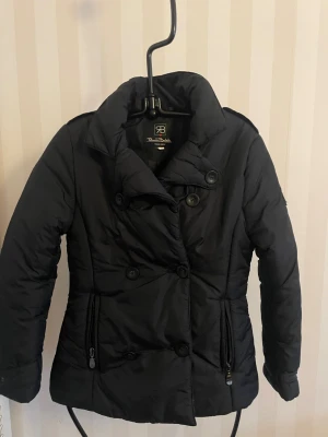 Svart pufferjacka från Renato Balestra - Svart pufferjacka med dubbelknäppning, höga krage och bälte i midjan från Renato Balestra.Jackan har dragkedjor på fickorna, långa ärmar och en snygg, figurnära passform. Perfekt för kalla dagar med sin vadderade polyesterkänsla. Taliensk storlek L motsvarar EU storlek XS/S, mått över axlar 38 cm, armlängd 55 cm.