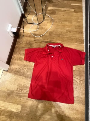 Röd Lacoste piké t-shirt - Säljer en röd piké t-shirt från Lacoste med klassisk krage och två knappar framtill. T-shirten har korta ärmar och den ikoniska krokodilloggan broderad på bröstet. Perfekt för dig som gillar sportig och stilren design.