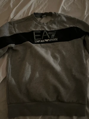 Grå sweatshirt från Emporio Armani - Säljer en stilren grå sweatshirt från Emporio Armani EA7 med svart panel och logga över bröstet. Tröjan har rund halsringning och långa ärmar. Perfekt för dig som gillar klassiska streetwear-vibbar.
