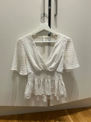 Vit blus med prickigt mönster och volang - Säljer en söt vit blus med prickigt mönster, omlott-v-ringning och korta, vida ärmar. Blusen har smock i midjan och volang nedtill som ger en flowy känsla. Perfekt för dig som gillar romantiska detaljer och ljusa toner.