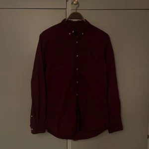 Ralph Lauren Skjorta - Snygg vinröd skjorta från Polo Ralph Lauren med klassisk button-down krage och broderad logga på bröstet. Skjortan har långa ärmar, vita knappar och är gjord i mjuk bomull. Perfekt för dig som gillar stilrena och tidlösa plagg.