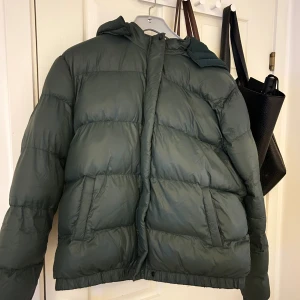 Mörkgrön pufferjacka med huva - Säljer en mörkgrön pufferjacka med stor, mysig huva och breda, quiltade sektioner. Jackan har dragkedja framtill och två sidofickor.  Jag köpte den förra vintersäsongen och den kom aldrig till användning. Därför så säljer jag den nu! 