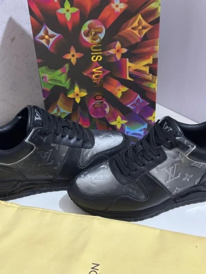 Louis Vuitton svarta sneakers i skinn - Snygga svarta sneakers från Louis Vuitton med silvergrå paneler och klassiskt LV-monogram över hela skon. Skorna har snörning, platt sula och är tillverkade i skinn med detaljer i syntet. Perfekta för dig som vill sticka ut med exklusiv stil. Storlek 41 men passar 42 lika så