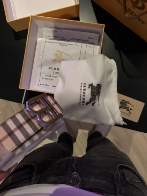 Burberry randigt skärp med B-spänne - Säljer ett Burberry bälte med klassiskt randigt mönster i beige, brun, vit och svart. Bältet har ett stilrent metallspänne med Burberrys B-logga och levereras med originalförpackning, dustbag och äkthetskort. Perfekt accessoar för att lyfta din outfit.