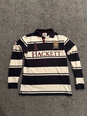 Randig pikétröja från Hackett London - Snygg långärmad pikétröja från Hackett London med breda marinblå och vita ränder, broderad logga och nummerdetalj på bröstet. Tröjan har klassisk krage, knappar och vinröda inslag vid halsen. Perfekt för dig som gillar sportig och preppy stil.