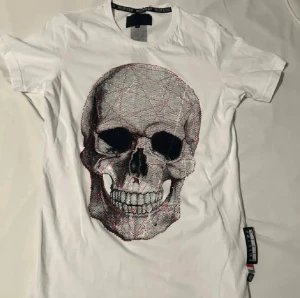 Philipp plein - Säljer en vit t-shirt från Philipp Plein med korta ärmar och rund hals. T-shirten har ett stort, glittrigt tryck framtill och coola detaljer på etiketten vid sidan. Perfekt för dig som gillar statement-plagg och exklusiv streetstyle.