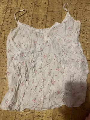 Söt blommig linnetopp med volang - Romantisk vit linnetopp med små rosa blommor och prickar. Toppen har smala axelband, dekorativa volanger framtill och tre pärlemorknappar vid halsringningen. Perfekt för en somrig look med vintagekänsla.