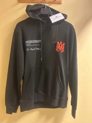 Svart AMIRI hoodie med tryck - Svart hoodie från AMIRI med stor huva och snörning. Framtill finns vit text och en röd AMIRI-logga, medan ryggen har ett stort tryck med text och koordinater i vitt och rött. Tröjan har en klassisk känguruficka och är gjord i 100% bomull. Perfekt för dig som gillar streetwear och snygga detaljer.