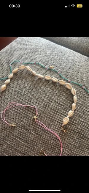 Sommarsmycken med snäckor och stjärna - Tre armband: ett med vita snäckor på beige tråd, ett med turkos och rosa pärlor samt liten snäcka, och ett med rosa tråd och guldfärgad sjöstjärna. Perfekta accessoarer för strandvibbar och sommarkänsla.