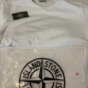 Vit långärmad tröja Stone Island - Vit långärmad tröja från Stone Island med klassisk rund hals och ikonisk svart-gul patch på vänster ärm. Tröjan är tillverkad i mjuk bomull och har en clean, stilren look som passar perfekt till jeans eller cargo. Märkeslogga i nacken och autenticitetslapp med QR-kod.