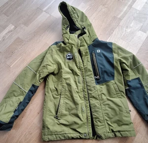 8848 Altitude Avanti JR Jacket - Snygg grön och mörkgrön vinter/skidjacka från 8848 Altitude med huva, flera dragkedjor och coola detaljer. Jackan har reflexdetaljer på ärmarna och praktiska fickor. Justerbar, avtagbar huva, specialutformad för hjälmen Heltejpade sömmar, för en helt vattentät design samt liftkortsficka på vänster ärm.