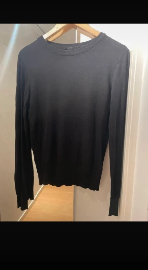 Svart stickad långärmad tröja - En stilren svart stickad tröja med rund hals och långa ärmar. Tröjan har en klassisk passform och är perfekt för dig som gillar enkla och snygga basplagg. Materialet är mjukt och skönt mot huden, och den passar till många olika outfits.