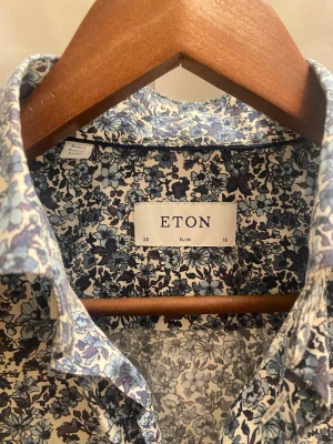 Blommig slim fit skjorta från Eton - Snygg skjorta från Eton med blommigt mönster i blå och lila toner på vit botten. Slim fit-modell med klassisk krage och knappar framtill. Tillverkad i mjuk bomull som känns skön mot huden. Perfekt för dig som gillar detaljer och vill sticka ut lite extra. Pris kan diskuteras vid snabb och smidig affär. Perfekt julklapp 🎄🎁.  Hör av er vid minsta frågan