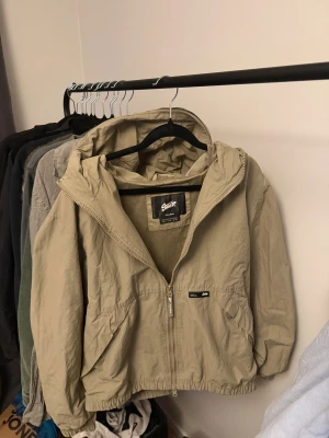 Grön  vindjacka från Pull&Bear - Snygg grön vindjacka från Pull&Bear med huva och dragkedja framtill. Jackan har två fickor, elastisk nederkant och meshfoder på insidan. Perfekt för blåsiga dagar och enkel att matcha med det mesta.