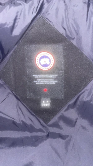 Svart pufferjacka från Canada Goose - Säljer en svart pufferjacka från Canada Goose med huva och klassisk logga på ärmen. Jackan har dragkedja framtill, två stora fickor och quiltad design. Perfekt för kalla dagar och riktigt snygg till streetwear-stilen.