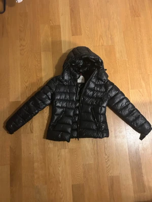 Svart Moncler dunjacka med huva - Säljer en svart dunjacka från Moncler med huva och tvåvägsdragkedja. Jackan har quiltad design, Moncler-logga på ärmen och praktiska fickor med dragkedja. Perfekt för kalla dagar och har en snygg, puffig look som är riktigt trendig.