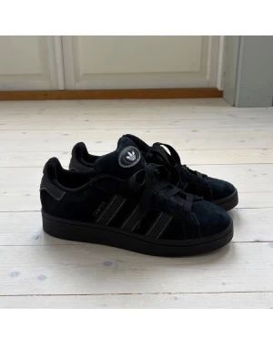 Svarta Adidas Campus sneakers mocka - Adidas Campus sneakers i svart mocka med klassiska tre ränder på sidan och snörning. Låg modell med rund tå och platt sula. Adidas-logga på plösen och hälen. Snygg och stilren design som passar till streetwear och casual outfits.
