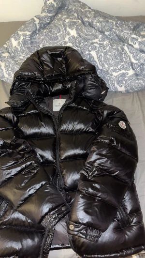 Moncler jacka - Säljer en svart pufferjacka från Moncler med glansig finish och stor huva. Jackan har dragkedja framtill, elastiska muddar och Moncler-logga på ärmen. Insidan har coolt serietryck och snörning vid nederkant. Perfekt för dig som vill ha en varm och snygg vinterjacka.