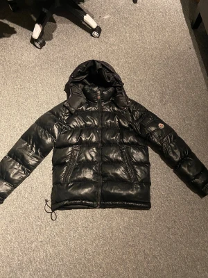 Svart Moncler pufferjacka med huva - Svart pufferjacka från Moncler med glansig finish, huva och dragkedja framtill. Jackan har två snedställda fickor med dragkedja och Moncler-logga på ärmen. Fylld med dun och har justerbar snörning nedtill. Perfekt för kalla dagar.
