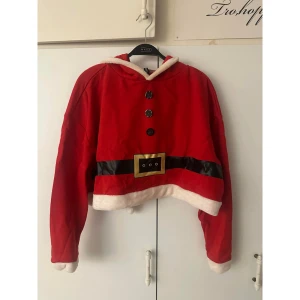 Röd julhoodie från H&M Divided - Säljer en röd hoodie (magtröja) med långa tomteluva från H&M Divided, inspirerad av tomtedräkt. Tröjan har svarta och vita detaljer, dekorativa knappar framtill, bälte-tryck och fluffig vit kant vid huva och nederkant. Perfekt för julmys och roliga bilder!
