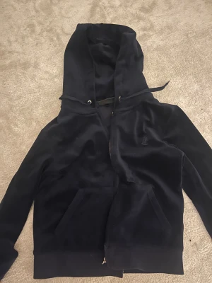 Mörkblå velour hoodie Juicy Couture - Säljer en mörkblå hoodie från Juicy Couture i mjuk velour med huva och snörning. Tröjan har en snygg dragkedja med logodetalj, känguruficka och broderad JC-logga på bröstet. Perfekt för chill dagar,streetstyle och speciellt nu till vintern. J-et på kedjan har gått sönder