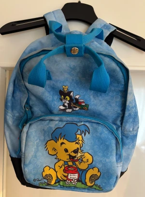 Blå Bamse ryggsäck med tryck - Söt blå ryggsäck med Bamse och hans vänner på framsidan. Väskan har justerbara axelremmar, ett stort fack och en mindre ficka med dragkedja. Materialet är slitstark polyester och designen är lekfull med tydliga figurer och färgglada detaljer.