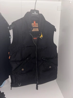 Svart dunväst från Parajumpers - Svart dunväst från Parajumpers med hög krage, dragkedja framtill och två snedställda fickor med dragkedja och knapp. Västen har detaljer i orange och gul samt broderad logga på ryggen. Perfekt för kyliga dagar. Perfekt skick haft den i max ett år, pris går att diskutera. 