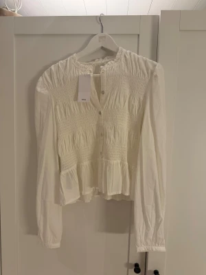 Vit smockad blus från MNG - Supersöt vit blus från MNG med smockad överdel och volang nedtill. Blusen har rundad hals, knappar framtill och långa ärmar med lätt puff. Materialet är mjukt och luftigt, perfekt för en chill och trendig look.Storlek barn:M men motsvarar S