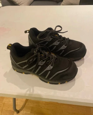 Svarta sneakers från Bluewear - Säljer ett par svarta Stålhetta skor från Bluewear med grå och gula detaljer. Skorna har snörning, mesh- och syntetmaterial samt robust sula med gula inslag. Perfekta för dig som vill ha en sportig och bekväm look på jobbet! Storlek 36 