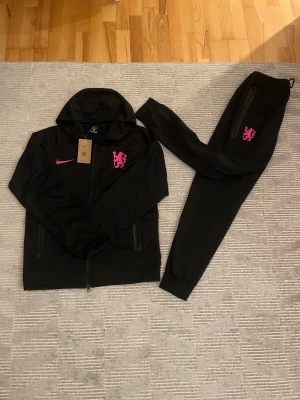 Svarta Chelsea nike tech Size M  - Svarta mjukisbyxor från Nike med rosa Swoosh och rosa lejonemblem. Byxorna har smal passform, ribbade muddar vid benslut och dragkedjeficka på sidan. Materialet är mjukt och följsamt, perfekt för chill eller träning. Snygg och sportig look med coola detaljer.