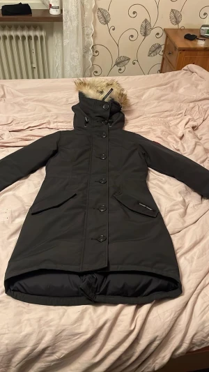 Svart rossclair parka jacka Canada Goose  - Säljer en svart dunjacka från Canada Goose med päls på huvan och hög krage. Jackan har långa ärmar, stora fickor med lock, knappar framtill och dragkedja bak. Perfekt för kalla dagar med snygg och stilren look. Finns kvitto  från johnells