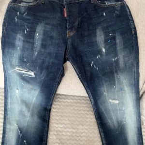 Dsquared2 blå distressed jeans, stl 33 - Säljer ett par blå jeans från Dsquared2 i storlek 33. Jeansen har en cool distressed look med slitningar, färgstänk och ljusa partier framtill och baktill. Klassisk femficksmodell med Dsquared2-logga bak i midjan. Perfekta för en edgy streetstil.